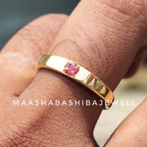 Puede incluir: Un anillo dorado con una pequeña gema de rubí de corte cuadrado engastada en el centro. El anillo es liso y pulido, con el texto "MAASHABASHIBAJEWELL" visible en la banda.