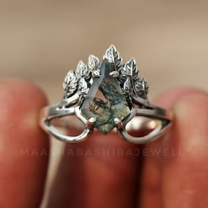 May include: A silver ring with a unique design. The ring features a central, faceted gemstone with green and brown inclusions, surrounded by leaf-shaped accents. The ring's band has an open design. The text "MAASHABASHIBAJEWELL" is visible.