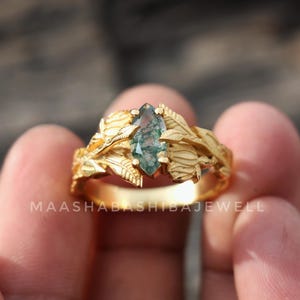 May include: A gold-colored ring with a marquise-cut, green gemstone. The ring features a leaf design around the gemstone and band. The text "MAASHABASHIBABJEWELL" is visible on the ring.