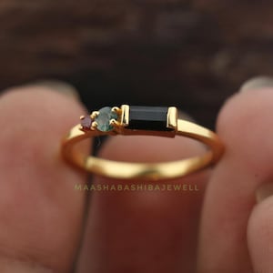 Può includere: Un anello d'oro con tre pietre preziose. L'anello presenta una pietra nera rettangolare affiancata da una piccola pietra viola e una pietra verde acqua. L'anello è tenuto da una persona. Il testo "MAASHABASHIBAJEWELL" è visibile.