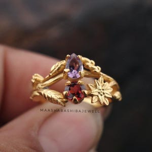 Può includere: Un anello color oro con un design floreale, con due pietre preziose: una pietra viola a forma di goccia e una pietra rossa a forma di cuore. L'anello ha un'estetica delicata ispirata alla natura.