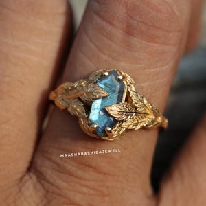 Może przedstawiać: Złoty pierścionek z prostokątnym, niebieskim kamieniem szlachetnym. Pierścionek zdobiony jest wzorem liści wokół kamienia. Na pierścionku widoczny jest napis "MAASHABASHIBAJEWELL".