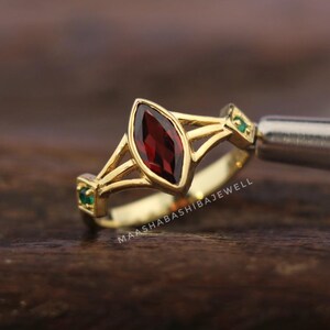 Puede incluir: Un anillo de oro con una piedra preciosa roja oscura en forma de marquesa como pieza central. El anillo presenta un diseño de caña dividida y está adornado con pequeñas piedras preciosas verdes a cada lado. El texto "MAASHABASHIBAJEWELL" es visible.