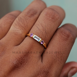 Op de afbeelding: Een gouden ring met een lichtblauwe rechthoekige edelsteen en twee kleinere paarse edelstenen aan weerszijden.