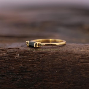 May include: A gold ring with a rectangular black gemstone. The ring is set on a wooden surface. The text "MAASHABASHIBAJEWEL" is visible on the wooden surface.