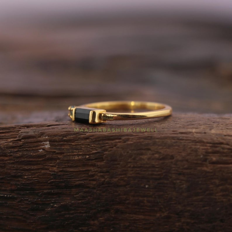 May include: A gold ring with a rectangular black gemstone. The ring is set on a wooden surface. The text "MAASHABASHIBAJEWEL" is visible on the wooden surface.