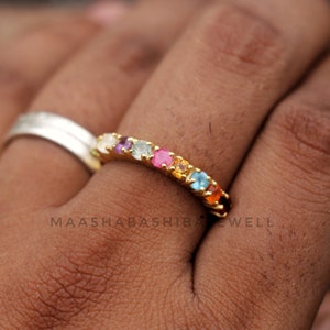 May include: A gold band ring with a row of colorful gemstones. The gemstones are set in a gold band and are arranged in a rainbow pattern. The ring is on a person's finger.