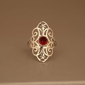 May include: A silver ring with a delicate filigree design and a red gemstone in the center.