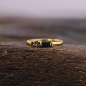 May include: A gold ring featuring a rectangular black gemstone flanked by a small red and green gemstone. The ring is set on a wooden surface. The ring is a piece of jewelry.