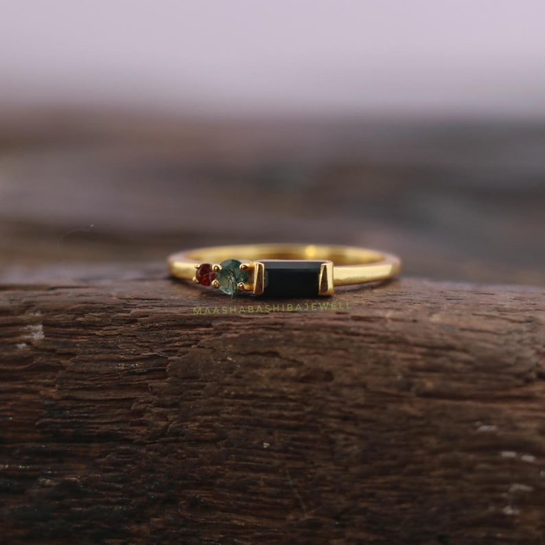 May include: A gold ring featuring a rectangular black gemstone flanked by a small red and green gemstone. The ring is set on a wooden surface. The ring is a piece of jewelry.