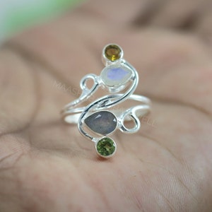 Könnte beinhalten: Ein silberner Ring mit einem filigranen, wirbelnden Design, das mit vier Edelsteinen verziert ist: einem hellblauen Mondstein, einem grauen Labradorit, einem grünen Peridot und einem braunen Citrin.