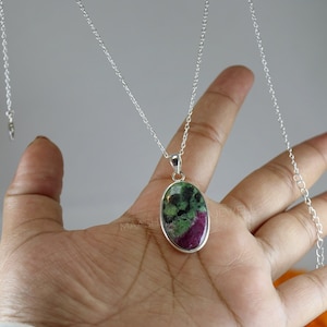 May include: A silver necklace featuring an oval pendant with a natural ruby zoisite stone. The stone displays a mix of green, pink, and black hues. The pendant is set in a silver bezel and hangs from a delicate silver chain.
