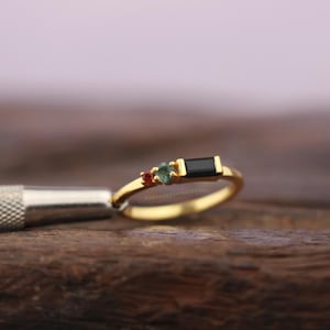 May include: A gold ring with three gemstones. The ring features a small red gemstone, a green gemstone, and a rectangular black gemstone. The ring is on a wooden surface.