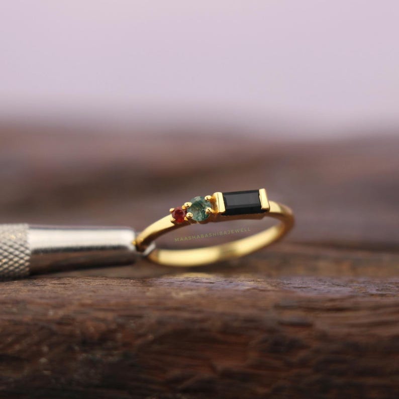 May include: A gold ring with three gemstones. The ring features a small red gemstone, a green gemstone, and a rectangular black gemstone. The ring is on a wooden surface.