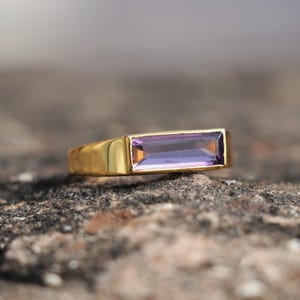 Puede incluir: Un anillo de oro con una piedra preciosa rectangular de color púrpura. El anillo tiene un diseño simple y moderno con un acabado pulido. La piedra preciosa es de color púrpura oscuro y está engastada en una banda de oro.