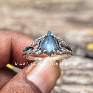 Puede incluir: Anillo de plata con una piedra preciosa azul en forma de cometa. El anillo tiene un diseño detallado en forma de hoja alrededor de la piedra. El texto "MAASHABASHIBAJEWELL" es visible.