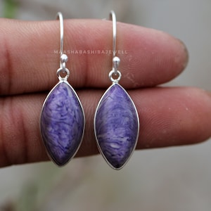 Puede incluir: Un par de pendientes de plata con gotas de piedra marmoleada de color morado y blanco. Los pendientes tienen un diseño simple de gancho.