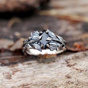 May include: A silver ring with a black gemstone surrounded by intricate floral designs. The ring is set on a wooden surface.