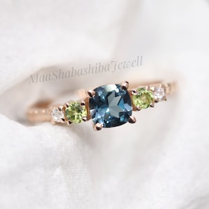 May include: Rose gold ring with a large, square, teal gemstone at its centre, complemented by smaller green and white stones. The band has a textured pattern. The ring is displayed against a soft, white backdrop.
