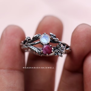 Op de afbeelding: Een zilveren ring met een delicaat bloemmotief met een maansteen en een robijn. De ring zit op een vinger.