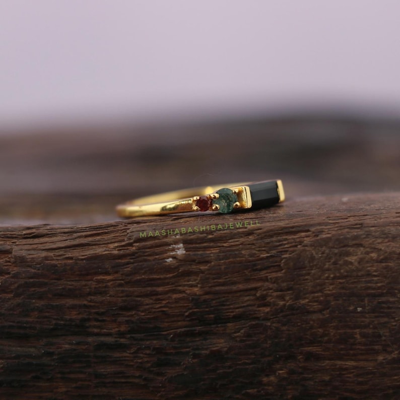 May include: A gold ring with a rectangular black stone and two smaller gemstones. The ring has a slender band and is set on a wooden surface. The text "MAASHABASHIBEJEWEL" is visible on the ring.