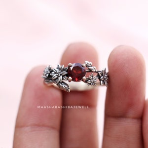 Puede incluir: Un anillo de plata con una piedra preciosa roja rodeada de delicadas flores de plata. El anillo está en un dedo.