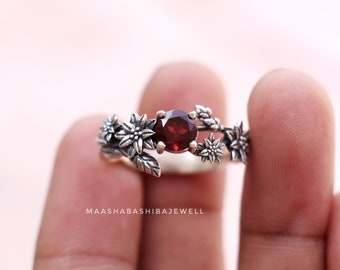 Puede incluir: Un anillo de plata con una piedra preciosa roja rodeada de delicadas flores de plata. El anillo está en un dedo.