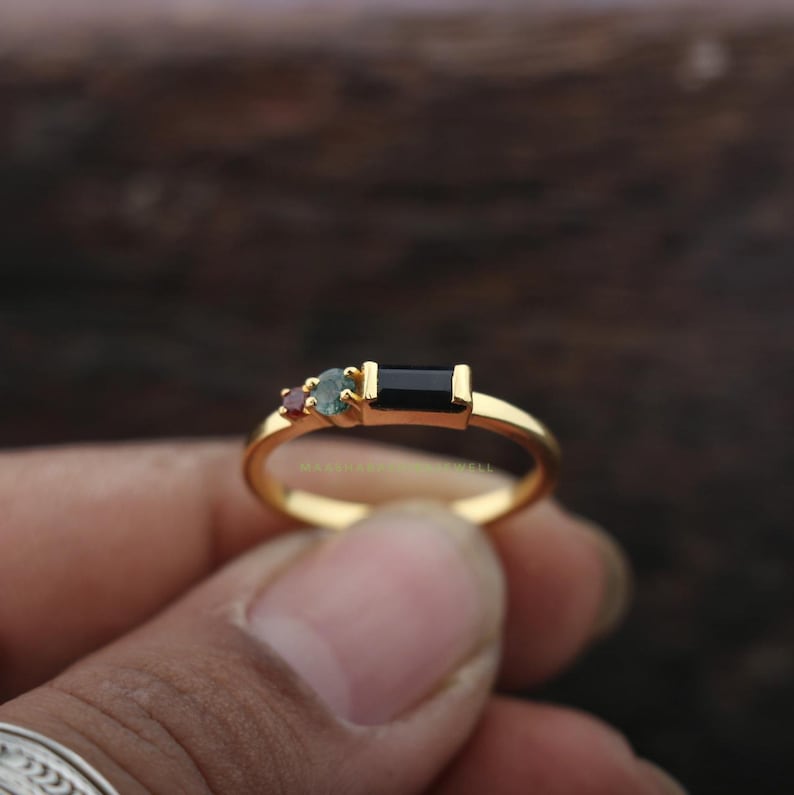 May include: A gold ring featuring three gemstones: a small red stone, a teal stone, and a rectangular black stone. The ring is held in a hand, showcasing the polished gold band and the arrangement of the stones.