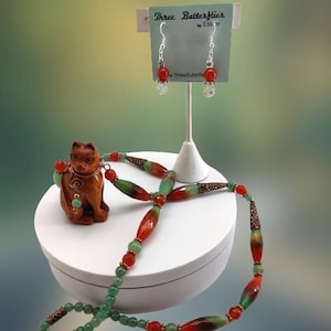 May include: A necklace and earrings set featuring green and orange gemstone beads. The necklace has a copper wire accent and the earrings are silver with orange gemstone drops. The set is displayed on a white stand with a wooden cat figurine.
