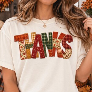Puede incluir: Una camiseta de manga corta color blanco roto con la palabra THANKS impresa en estilo patchwork. Cada letra está compuesta por diferentes patrones de tela, incluyendo cuadros, vichy y estampados florales en tonos naranja, rojo, verde y amarillo.