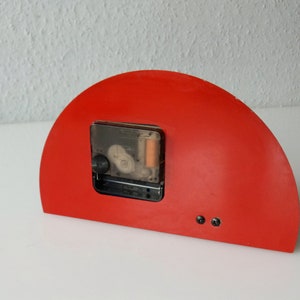May include: Red half-circle clock with a black clock face and a clear plastic cover. The clock is mounted on a white surface.