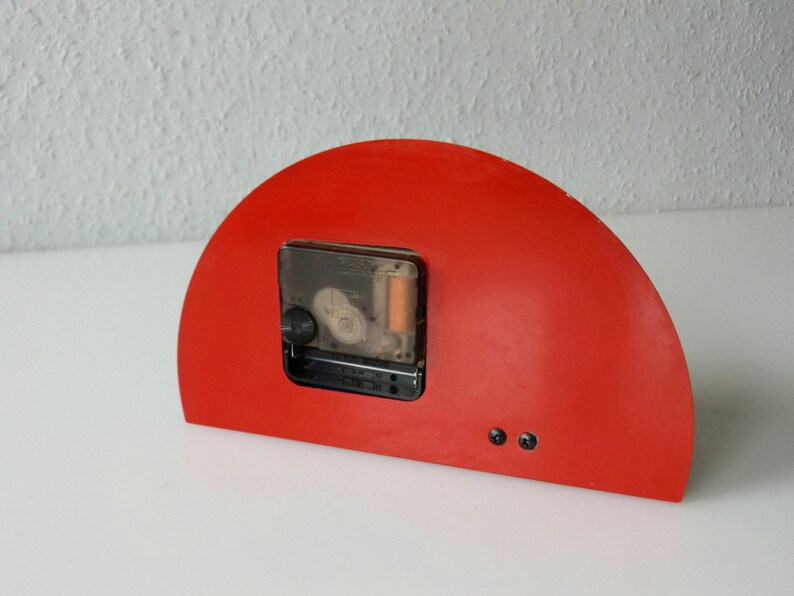 May include: Red half-circle clock with a black clock face and a clear plastic cover. The clock is mounted on a white surface.