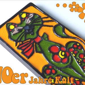 May include: A ceramic tile with a green cat design with red, yellow, and black accents. The cat is wearing a red bow and is surrounded by orange flowers. The tile has a black border and a yellow background. The text "70er Jahre Kult" is printed in orange at the bottom of the tile.
