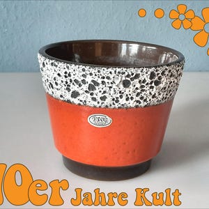 May include: A vintage ceramic planter with a textured orange body, a speckled white and black rim, and a dark brown interior. The planter has a small, oval sticker and the text "70er Jahre Kult" in orange.