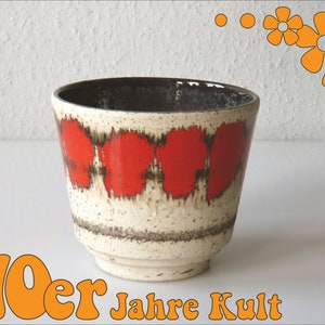 Può includere: Un vaso di ceramica bianco con un motivo geometrico rosso e marrone. Il vaso ha una base larga e un'apertura stretta. Il testo "70er Jahre Kult" è stampato sotto il vaso.