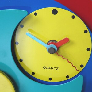 May include: A colorful clock with a yellow face, blue and green outer rings, and red and blue hands. The clock face has black numbers and the word "QUARTZ" printed on it.