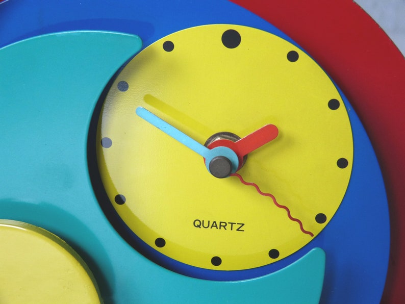 May include: A colorful clock with a yellow face, blue and green outer rings, and red and blue hands. The clock face has black numbers and the word "QUARTZ" printed on it.