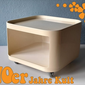 Könnte beinhalten: Ein beigefarbener, quadratischer Beistelltisch mit flacher Oberseite und offenem Stauraum. Der Tisch hat abgerundete Ecken und steht auf kleinen Rollen. Der Text "70er Jahre Kult" ist unten im Bild orange dargestellt.