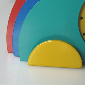 May include: A colorful clock with a yellow face, blue and red accents, and a yellow base. The clock is made of wood and has a modern design.