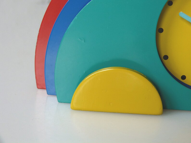 May include: A colorful clock with a yellow face, blue and red accents, and a yellow base. The clock is made of wood and has a modern design.