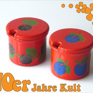 Può includere: Due zuccheriere in ceramica rossa con coperchi. Una ciotola ha un design di more nere e verdi. L'altra ciotola ha un design di prugne blu e verdi. Il testo "70er Jahre Kult" è stampato sotto le ciotole.