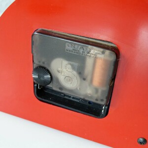 May include: Close-up of the back of a red clock with a clear quartz movement. The clock has a black knob for setting the time and the text "QUARTZ MADE IN CHINA" is visible on the movement.