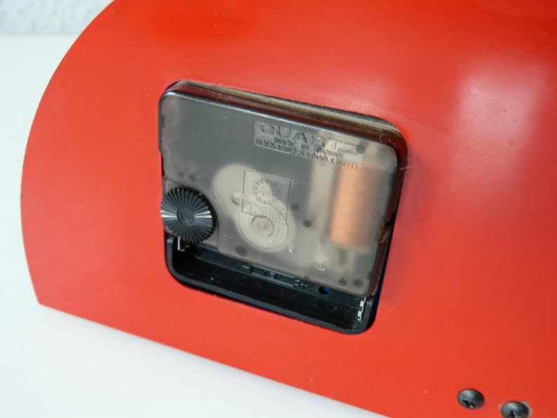 May include: Close-up of the back of a red clock with a clear quartz movement. The clock has a black knob for setting the time and the text "QUARTZ MADE IN CHINA" is visible on the movement.