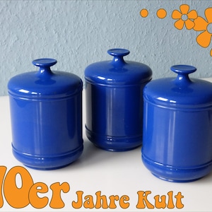 Peut inclure: Trois pots en céramique bleus avec couvercles. Les pots sont ronds et ont une finition lisse et brillante. Les couvercles sont légèrement bombés et ont un petit bouton sur le dessus. Les pots sont disposés en rangée sur une surface blanche. Le texte "70er Jahre Kult" est visible dans la partie inférieure de l'image.