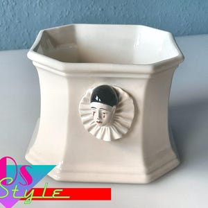 Puede incluir: Jardinera de cerámica color crema con forma hexagonal. Presenta un relieve decorativo de una cara de payaso llorando con sombrero negro y cuello con volantes. La jardinera tiene una amplia abertura.