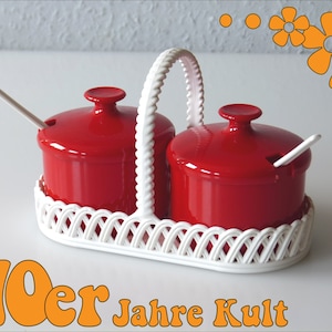 Può includere: Un vassoio da portata in ceramica bianca con un design intrecciato contiene due barattoli per condimenti in ceramica rossa con coperchi e cucchiai. I barattoli hanno una piccola apertura sul coperchio per il cucchiaio.