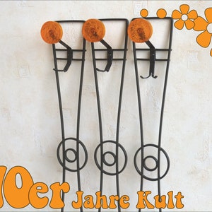 Könnte beinhalten: Ein schwarzer Metall-Kleiderständer mit drei Haken und orangefarbenen runden Akzenten. Der Ständer ist an einer weißen Wand mit einem Muster aus kleinen Quadraten montiert. Der Text "70er Jahre Kult" ist in orangefarbenen Buchstaben am unteren Rand des Bildes geschrieben.