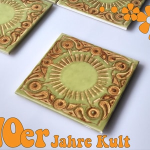 May include: Square ceramic tiles featuring a light green glaze with a central sunburst motif surrounded by brown embossed floral patterns. The image includes orange graphic flower icons and the text 70er Jahre Kult in orange font.