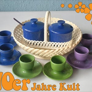Può includere: Un set di sei portauova in ceramica con piattini abbinati in blu, verde e viola, disposti attorno a un cestino in plastica color crema con manico centrale. Due contenitori blu con coperchio e un cucchiaino sono all'interno. Testo 70er Jahre Kult.
