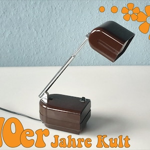 Könnte beinhalten: Eine braune und schwarze Schreibtischlampe mit einem Chromarm. Die Lampe hat einen rechteckigen Sockel und einen geschwungenen Schirm. Das Bild enthält den Text "70er Jahre Kult" in Orange sowie orangefarbene Blumengrafiken.